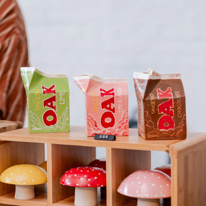 Three OAK branded ceramic milk cartons on a wooden shelf with decorative mushrooms below.